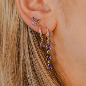 Può includere: Orecchini dorati con gemme viola a forma di goccia. L'immagine mostra un orecchino a farfalla e un design a cerchio pendente. I gioielli sono ambientati su uno sfondo di capelli biondi e pelle, evidenziando il design e il colore.