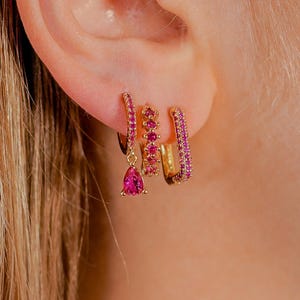 Może przedstawiać: Złote kolczyki z różowymi kamieniami szlachetnymi. Zestaw zawiera kolczyk z kroplą, małe kółko z rzędem kamieni i większe kółko z pełnym rzędem różowych kamieni.