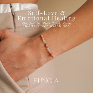 May include: A delicate gold bracelet adorned with pink, green, and gold beads. The bracelet is displayed on a wrist, with the text "Self-Love & Emotional Healing" and "EUNOIA" visible in the background. The bracelet features Rhodonite, Pink Opal, Rose Quartz, and Green Aventurine.