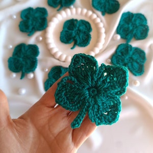 May include: A set of eight crocheted green four-leaf clover charms. The charms are arranged on a white surface with white pearls.
