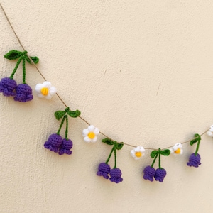 Puede incluir: Una guirnalda de ganchillo hecha a mano con flores moradas en forma de campana y margaritas blancas con centros amarillos, ensartadas en hilo natural. Un elemento decorativo para un toque caprichoso.