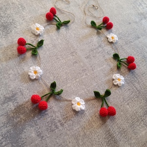 Puede incluir: Una guirnalda de crochet con margaritas blancas y cerezas rojas. La guirnalda está enhebrada en un cordel natural.
