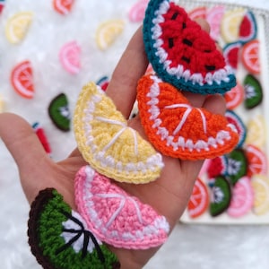 Könnte beinhalten: Eine Sammlung gehäkelter Fruchtscheiben, darunter Wassermelone, Orange, Zitrone, rosa Grapefruit und Kiwi. Die Fruchtscheiben befinden sich alle in einer Hand, und im Hintergrund befinden sich weitere Fruchtscheiben.