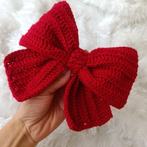 Könnte beinhalten: Eine große rote gehäkelte Schleife. Die Schleife ist aus einem einzigen Garnfaden gefertigt und hat ein schlichtes, klassisches Design.