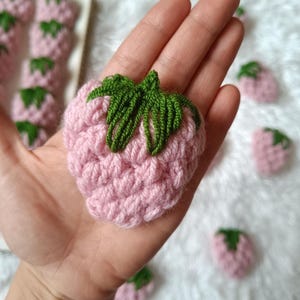 Peut inclure: Gros plan sur une fraise en crochet rose avec des feuilles en crochet vert. La fraise est tenue dans la main d'une personne. Il y a d'autres fraises en crochet en arrière-plan.