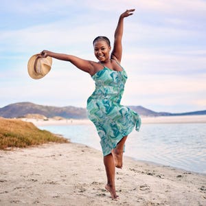 May include: A woman in a teal and white patterned, sleeveless dress, holding a straw hat, on a sandy beach. The dress has a flowing design. The background shows a sea, mountains, and a bright sky.