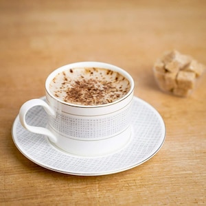 Op de afbeelding: Een witte keramische koffiekop met een grijs en wit patroon, gevuld met koffie en bedekt met een laagje kaneel, op een bijpassend schoteltje.