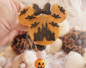 May include: A Halloween-themed Mickey Mouse-shaped badge holder with a black castle silhouette, bats, and a glitter finish. The holder has a black and orange pumpkin charm and a clear plastic clip.