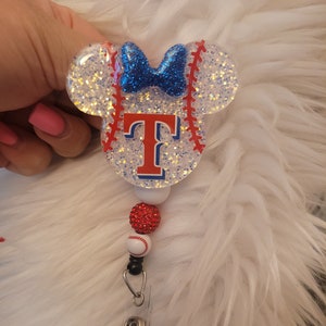 May include: A white and red baseball-themed Mickey Mouse-shaped badge holder with a blue bow and the letter "T" in red and blue. The badge holder is attached to a red and white baseball bead and a clear plastic badge clip.