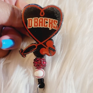 May include: A black heart-shaped badge holder with the text "D-BACKS" in red and white. The badge holder is attached to a retractable badge reel with a baseball charm and red rhinestone beads.