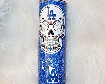 May include: A blue glitter tumbler featuring a sugar skull design with baseballs for eyes and the Los Angeles Dodgers logo. The tumbler also displays the team's logo with a palm tree. The tumbler is approximately 8 inches tall.