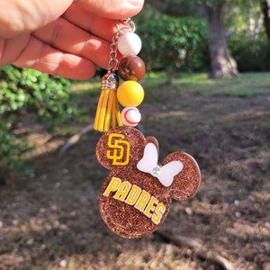 May include: A brown glitter keychain featuring a mouse-shaped design with the text "PADRES" in yellow. The keychain includes a yellow tassel, beads, and a baseball charm. The San Diego Padres logo is visible.