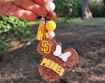 May include: A brown glitter keychain featuring a mouse-shaped design with the text "PADRES" in yellow. The keychain includes a yellow tassel, beads, and a baseball charm. The San Diego Padres logo is visible.