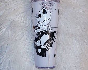 May include: A white and black tumbler with a graphic of Jack Skellington and Zero from the Nightmare Before Christmas. The tumbler has a purple lid and a clear straw.