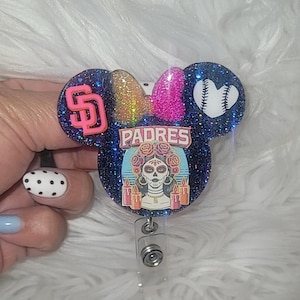May include: A glittery badge reel in the shape of Mickey Mouse ears. The design features the San Diego Padres logo, a baseball, and a Day of the Dead sugar skull. The bow is pink and gold. The word "PADRES" is displayed.
