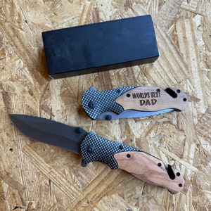May include: A black pocket knife with a wood handle and a carbon fiber pattern on the blade. The knife has a safety latch and a belt clip. The handle is engraved with the words "World's Best Dad".