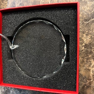 May include: A clear, faceted glass ornament with a circular shape, presented in a red box with black foam padding. A silver string is attached for hanging. The ornament's edges are cut with multiple facets.