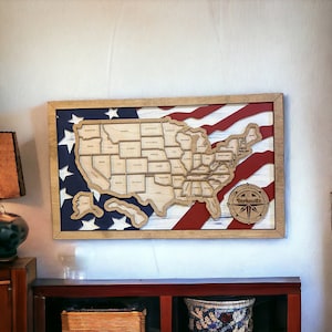 May include: A wooden map of the United States with each state outlined and labeled. The map is mounted on a red, white, and blue American flag background. The map is framed in a light brown wood frame.