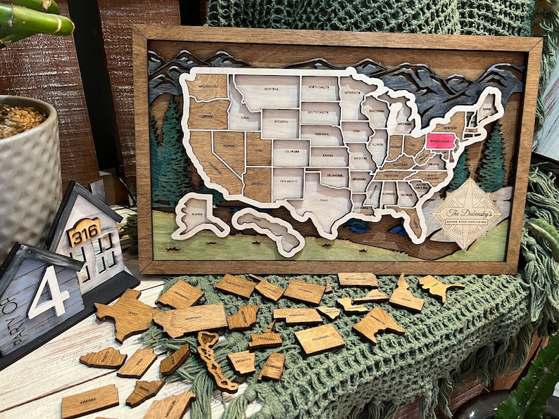 Puede incluir: Un mapa de madera de los Estados Unidos con cada estado como una pieza separada. El mapa est&aacute; enmarcado en un marco de madera oscura y tiene una escena de monta&ntilde;a en el fondo. Los estados est&aacute;n todos en sus posiciones correctas y est&aacute;n listos para ser colocados en el mapa. El texto "The Doleskey's" est&aacute; en el mapa.