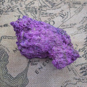 May include: A raw, jagged mineral specimen with a vibrant, deep purple crystalline texture. The surface of the stone is uneven and porous, featuring small, sparkling facets that catch the light, creating a rich, saturated colour throughout the piece.