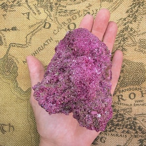 May include: A raw, crystalline mineral specimen with a vibrant magenta and deep purple coloration. The surface features a rough, jagged texture with numerous small, reflective facets that catch the light, creating a sparkling appearance.