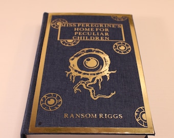Op de afbeelding: Een donkerblauw hardback boek met een gouden reliëftitel "Miss Peregrine's Home for Peculiar Children" en auteur "Ransom Riggs". De omslag heeft een gouden illustratie van een oog met een draaiend patroon.