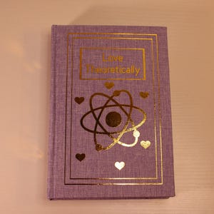 Op de afbeelding: Een paars hardback dagboek met een gouden ontwerp. De titel van het dagboek is "Love Theoretically" en bevat een gouden atoom met hartjes eromheen.