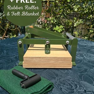 May include: A green metal printing press with a wooden base plate, accompanied by a black rubber roller and a folded green felt blanket. The image includes the text: + FREE: Rubber Roller & Felt Blanket.