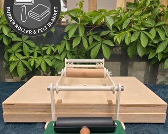 May include: A printmaking press featuring a light-colored metal frame and a wooden base. A black rubber roller with a wooden handle rests on a folded green felt blanket. A circular graphic includes the text GIFT RUBBER ROLLER & FELT BLANKET.
