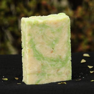 May include: A rectangular bar of handmade soap with a light green and cream swirl pattern. The soap is sitting on a black surface, with a blurred green and brown background.