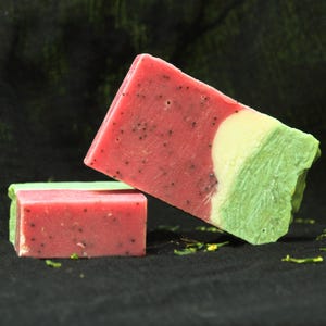 Könnte beinhalten: Zwei rechteckige Seifenstücke mit geschichtetem Design. Die Seife hat einen rosafarbenen Bereich mit kleinen schwarzen Punkten, einen dünnen weißen Streifen und einen hellgrünen Bereich. Die Stücke sind so angeordnet, dass eines steht und eines liegt.