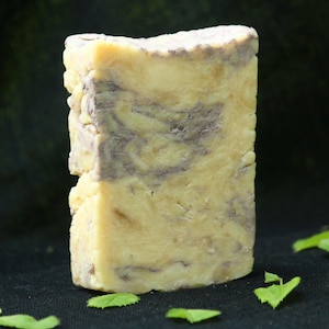 May include: A rectangular bar of soap with a marbled texture featuring creamy yellow and muted purple swirls. The surface of the soap appears solid and dense with a slightly uneven, hand-cut edge along the top, showcasing a natural color blend.
