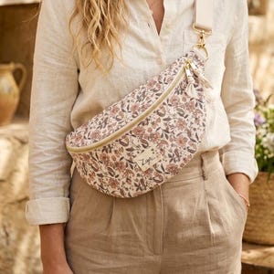 May include: A floral print bum bag with a cream-coloured strap and gold-tone hardware. The bag features a zip closure and a small tassel. The floral pattern includes shades of pink, brown, and cream. The bag is worn across the body.