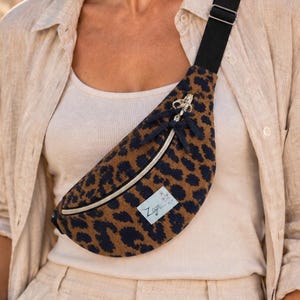 May include: A crescent-shaped fanny pack featuring a brown and black leopard print pattern. The bag has a black adjustable strap, a silver zipper closure with a dark ribbon pull, and a small rectangular white label with the text Zige.
