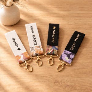 Peut inclure: Quatre porte-clés en tissu avec des mousquetons en métal doré. Deux sont blanc cassé avec des motifs floraux et le texte Mamie et SUPER MAMIE. Deux sont noirs avec des motifs floraux et le texte Best Gramy et Super Mamie avec un cœur.
