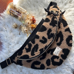 Puede incluir: Un bolso de cintura con estampado de leopardo beige y negro con una correa y cremallera negras. El bolso tiene una pequeña etiqueta con texto y está decorado con lazos negros. El bolso está sobre una superficie blanca y esponjosa.