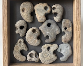 May include: A square light-colored wooden frame containing eleven natural gray and tan stones arranged on a dark gray fabric backing. Each stone features a distinct, naturally occurring hole or indentation, creating a collection of unique, textured, and weathered geological specimens displayed within the shadow box.