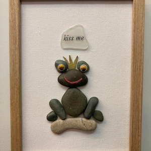 May include: A framed artwork featuring a frog made of stones. The frog is sitting on a stone and has a crown on its head. The frog has googly eyes and a red smile. The artwork has a white background and a wooden frame. The text "kiss me" is written on a white stone above the frog.