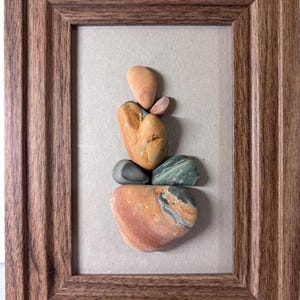 Puede incluir: Una obra de arte enmarcada con una disposición equilibrada de piedras de colores. Las piedras, en tonos naranja, verde y gris, están apiladas para parecerse a una figura. El marco es de madera con un acabado marrón cálido, lo que añade una estética natural a la pieza.