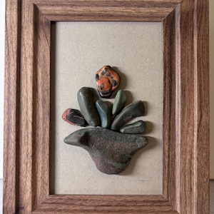 Puede incluir: Una obra de arte enmarcada con una disposición de piedras. La disposición incluye piedras verdes, marrones y naranjas de varios tamaños. El marco de madera tiene un acabado natural y la obra de arte está montada sobre un fondo neutro.