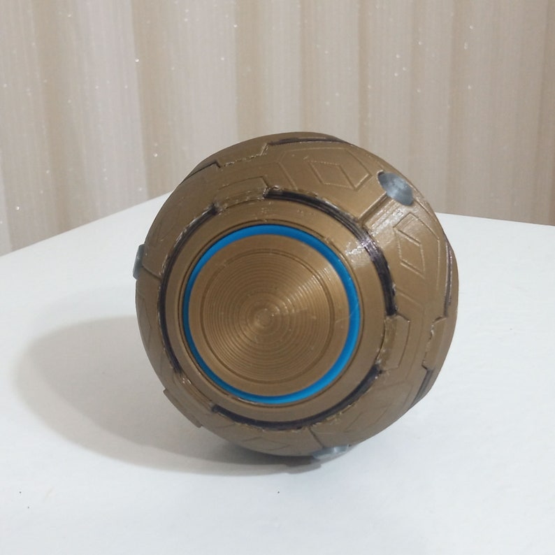 May include: A spherical, metallic-looking object with a complex geometric design. The object is primarily gold with a blue circular accent in the center. It has a textured surface and appears to be made of multiple connected panels.