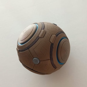 May include: A brown, spherical object with a geometric design. The sphere features circular and rectangular patterns, with accents of blue and gray. The design includes concentric circles and small, circular gray buttons. The object appears to be 3D-printed.
