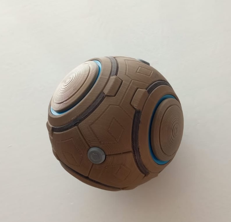 May include: A brown, spherical object with a geometric design. The sphere features circular and rectangular patterns, with accents of blue and gray. The design includes concentric circles and small, circular gray buttons. The object appears to be 3D-printed.