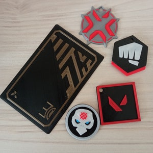 May include: Five 3D printed keychains featuring different designs. One keychain is a black and gold card with a geometric pattern. Another keychain is a gray and red circular design with a starburst pattern. A third keychain is a black and red design with a fist. A fourth keychain is a black and red square with a stylized V. The fifth keychain is a black and white circular design with a skull and crossbones.