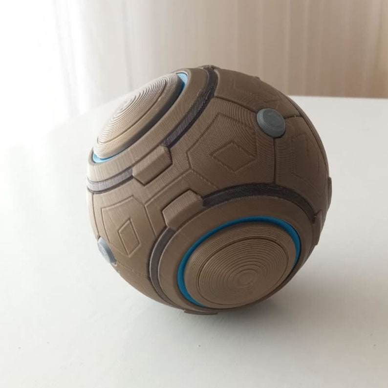 May include: A spherical, geometric object with a tan and gray color scheme. The ball features a complex pattern of raised lines, geometric shapes, and circular accents with a blue outline. Two small gray circular buttons are visible.