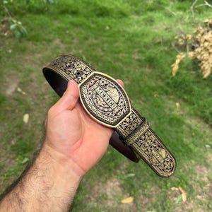 May include: Ornate gold-colored belt with a brown leather backing. The belt buckle features the word "MEXICO" and an emblem. The belt is decorated with detailed patterns and designs.