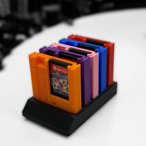 May include: A black stand holding several colorful Nintendo Switch game cartridges. The cartridges are in orange, red, pink, purple, and blue. One orange cartridge displays the game "Mario Kart".