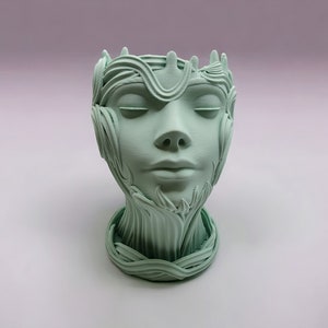 May include: A light green ceramic planter shaped like a woman's head with intricate details resembling leaves and vines.