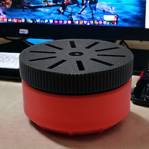May include: A red and black cylindrical container with a black lid. The lid has a circular design with multiple slots.