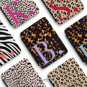 May include: A collection of rectangular protective cases featuring various patterns including leopard print, zebra stripes, and tortoiseshell. Several cases are personalized with large, bold, colored capital letters and a matching dot.
